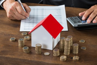 businessman calculating tax by model house and stacks of coins on table